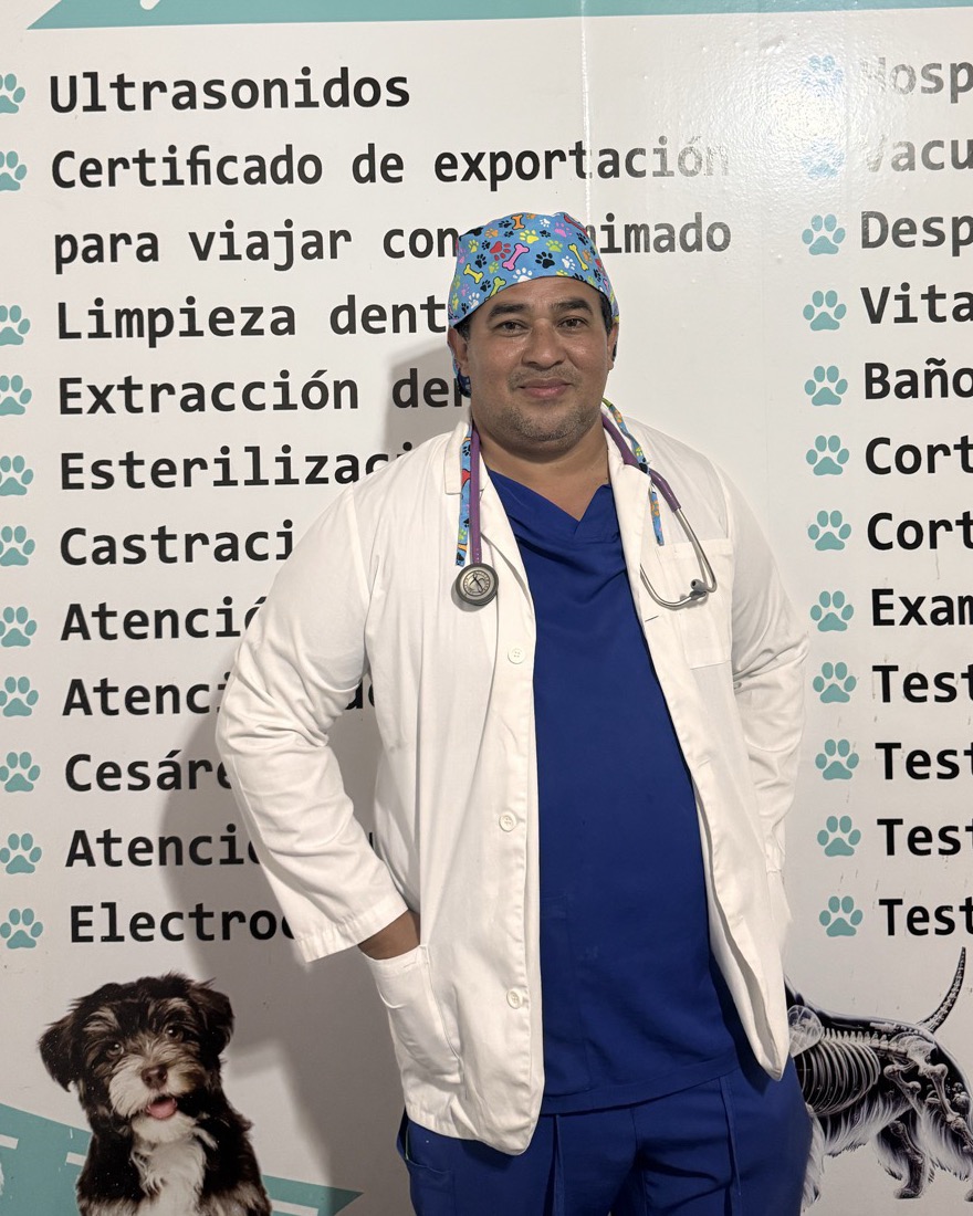 Dr. Ignacio Campos con gorro quirúrgico, bata blanca, uniforme azul y estetoscopio, de pie frente al rótulo de servicios de Centro Veterinario Los Mimados